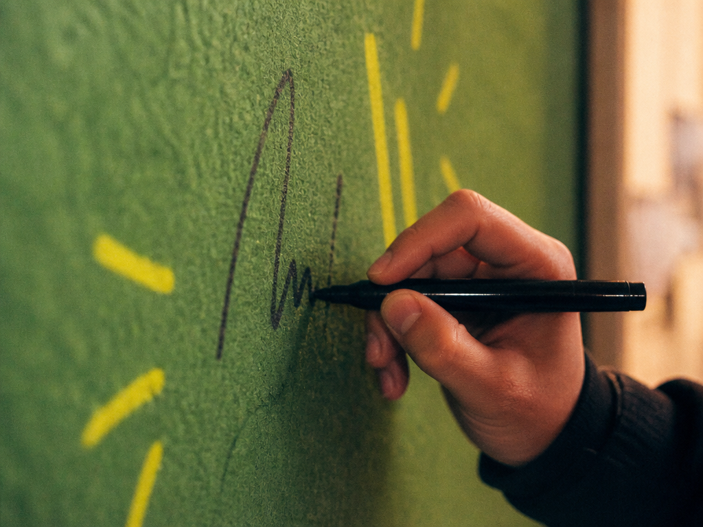 A hand writing a name on the felt-green clubhouse wall with a marker